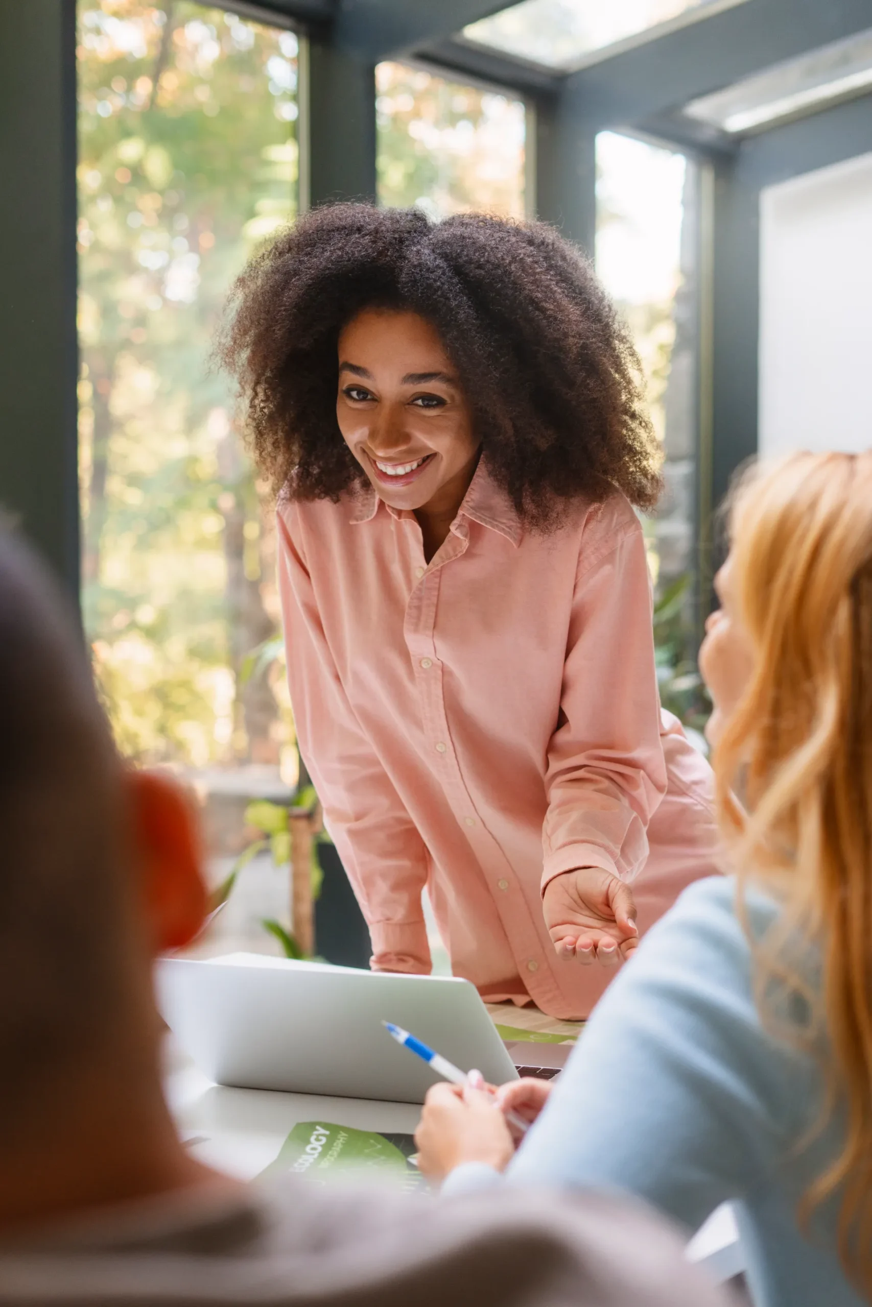 Femme souriante aux cheveux afro portant une chemise rose, discutant avec deux collègues autour d'un ordinateur portable.