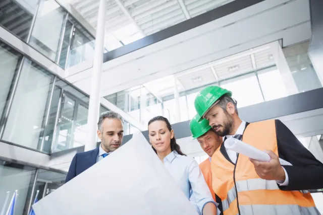 Quatre personnes, dont trois portant des casques de chantier, examinent des plans dans un bâtiment moderne.