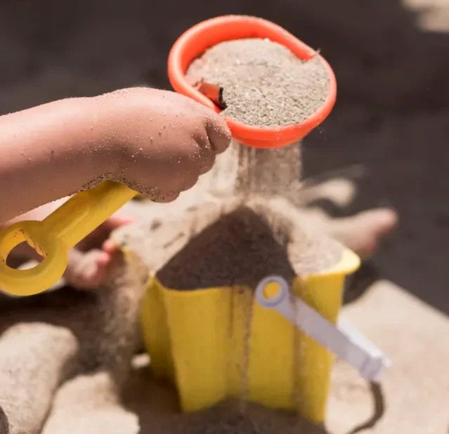 Une main d'enfant tient une pelle orange remplie de sable, versant du sable dans un seau jaune en forme d'étoile.