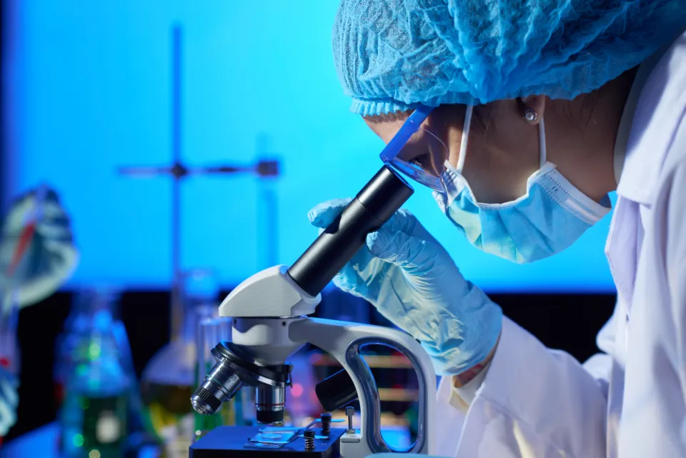Une scientifique portant une blouse, un masque et des gants observe une lame sous un microscope dans un laboratoire.